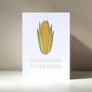 May include: A white card with a drawing of a yellow ear of corn with green husks. The text "Congratulations it's your birthday" is printed below the corn.