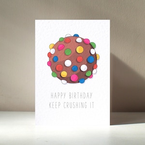Puede incluir: Tarjeta de felicitación blanca con una ilustración redonda marrón que se asemeja a una bola de pastel de chocolate cubierta de chispas de colores. El texto "HAPPY BIRTHDAY KEEP CRUSHING IT" está impreso en negro.