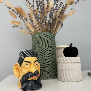 May include: A green vase with a leafy pattern holds a bouquet of dried lavender and pampas grass. A ceramic figurine of a man with a moustache and beard sits on a white surface next to a white ceramic jar with a geometric design.