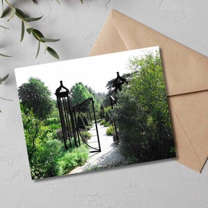 May include: A black metal garden gate with a curved design, surrounded by lush green foliage. The gate leads to a path through the garden.