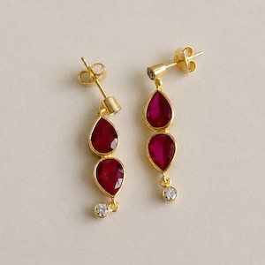 May include: A pair of gold drop earrings featuring two faceted, teardrop-shaped red stones suspended vertically. Each earring is topped with a small, clear, round-cut stone at the post and a second clear stone at the very bottom of the design.