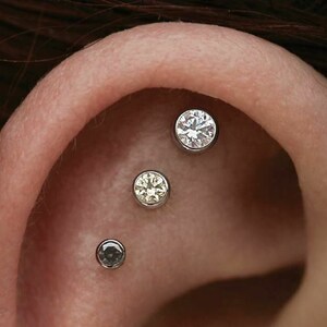 May include: Close-up of an ear with three piercings. The top piercing has a clear round gemstone, the middle piercing has a yellow round gemstone, and the bottom piercing has a black round gemstone.