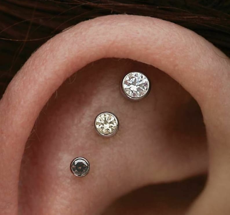 May include: Close-up of an ear with three piercings. The top piercing has a clear round gemstone, the middle piercing has a yellow round gemstone, and the bottom piercing has a black round gemstone.