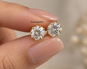 May include: Two round-cut clear stud earrings set in gold-toned metal with six-prong settings. The text Kweengems is visible above the earrings. Each earring features a faceted stone that reflects light, held securely by small metal prongs.