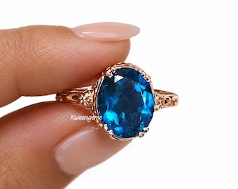 May include: A rose gold ring featuring a large, oval-cut, deep blue gemstone set in a prong setting. The band displays intricate, decorative metalwork along the sides. The text Kweengems is visible on the surface of the blue gemstone.