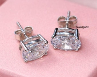 May include: A pair of silver-toned stud earrings featuring oval-cut clear stones held by four prongs. The earrings include standard butterfly backs and are displayed with the text Kweengems visible near the right earring on a pink surface.