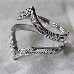 Può includere: Un anello color argento con un design a forma di V. L'anello presenta un motivo dettagliato di linee e curve, creando un aspetto strutturato. L'anello è mostrato su una superficie di colore chiaro.