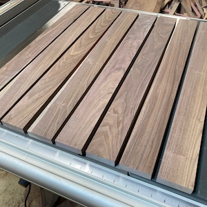 Könnte beinhalten: Acht Stücke Walnussholz, jedes etwa 45 cm lang, liegen auf einer Tischkreissäge. Das Holz ist hellbraun mit einer natürlichen Maserung.