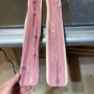 May include: Two unfinished cedar wood planks with a natural grain pattern and a reddish-purple hue. The planks have a curved shape and are ready for woodworking projects. The wood has knots and a smooth surface.