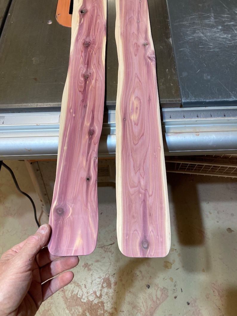 May include: Two unfinished cedar wood planks with a natural grain pattern and a reddish-purple hue. The planks have a curved shape and are ready for woodworking projects. The wood has knots and a smooth surface.