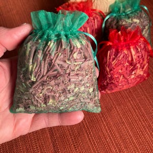 Könnte beinhalten: Eine Hand hält einen kleinen grünen Netzbeutel, der mit getrockneten Kräutern gefüllt ist. Weitere Beutel in Rot und Grün sind im Hintergrund zu sehen. Die Beutel sind mit getrocknetem, zerkleinertem Pflanzenmaterial gefüllt. Ein Blumenstrauß ist im Hintergrund.