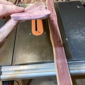 May include: A piece of unfinished cedar wood, held up to show the grain and color. The wood is a light purple-red hue. A long, unfinished cedar plank is next to it on a table saw.