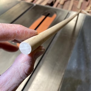 May include: A light-colored wooden dowel rod, held between fingers, with a rounded end. The dowel is smooth and cylindrical, likely for woodworking or craft projects. The background shows a saw and other wood pieces.