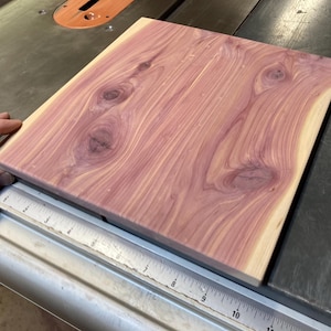 May include: A square piece of wood with a reddish-purple hue and a natural wood grain pattern. The wood has a knot in the center and is being measured with a ruler.