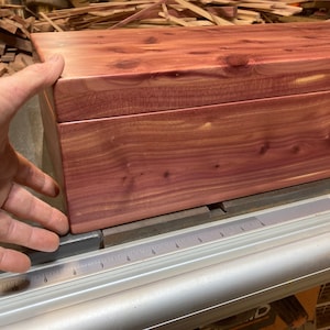 Può includere: Una scatola rettangolare in legno di cedro marrone rossiccio. La scatola è su una superficie metallica con un righello. La venatura del legno è visibile. La scatola è tenuta da una mano.