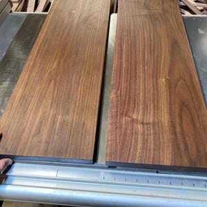 Könnte beinhalten: Zwei Stücke dunkelbraunes Holz, wahrscheinlich Walnuss, liegen nebeneinander auf einer Tischkreissäge. Das Holz ist glatt und hat eine natürliche Maserung.
