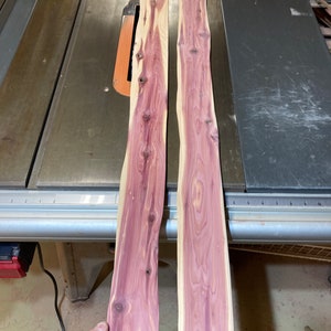 May include: Two unfinished cedar wood planks with a natural edge, displaying a vibrant reddish-purple hue with visible wood grain and knots. The planks are positioned on a metal table saw, with a pile of wood scraps in the background.