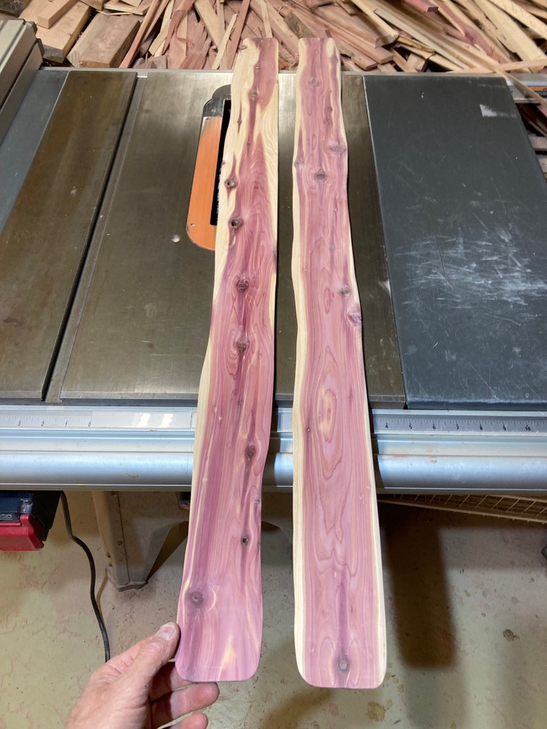 May include: Two unfinished cedar wood planks with a natural edge, displaying a vibrant reddish-purple hue with visible wood grain and knots. The planks are positioned on a metal table saw, with a pile of wood scraps in the background.