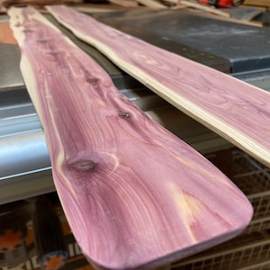May include: Two unfinished cedar wood planks with a natural purple and cream color. The planks have a smooth, sanded surface and visible wood grain. The planks are likely for woodworking projects.