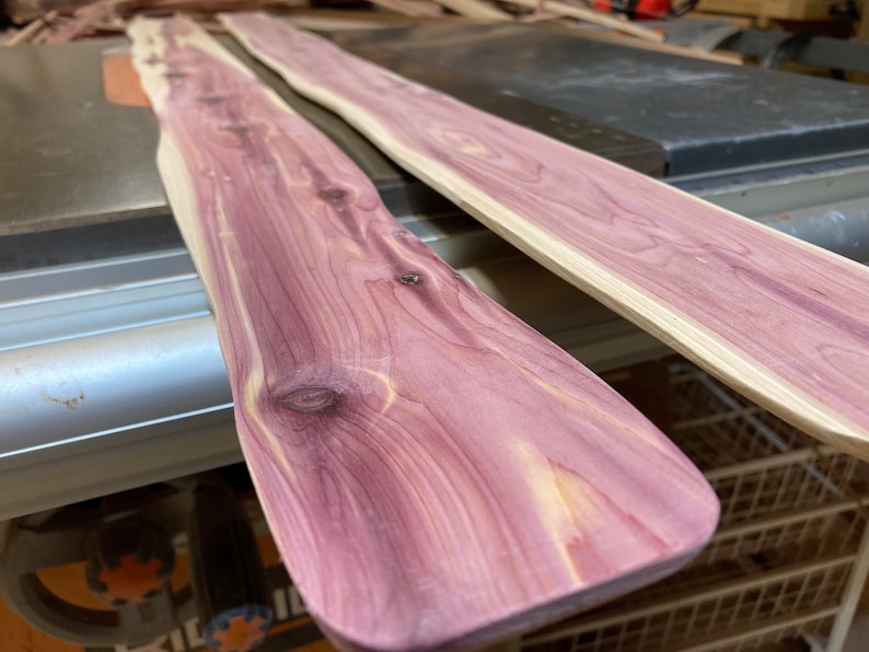 May include: Two unfinished cedar wood planks with a natural purple and cream color. The planks have a smooth, sanded surface and visible wood grain. The planks are likely for woodworking projects.