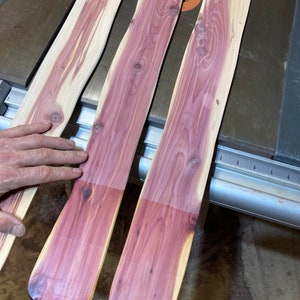 May include: Three unfinished wooden planks with a natural grain pattern. The planks have a light tan outer edge and a vibrant purple-red center. The wood is smooth and appears to be freshly cut, with a slight curve along the edges.