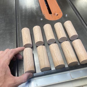 Op de afbeelding: Elf onafgewerkte houten deuvels, cilindrisch van vorm, gerangschikt op een metalen oppervlak. De deuvels zijn lichtbruin van kleur en lijken van hardhout te zijn. Eén hand houdt één van de deuvels vast.