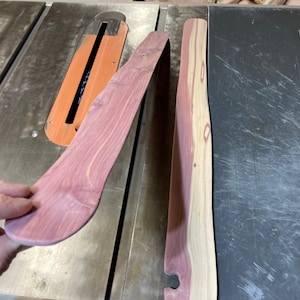 Puede incluir: Dos piezas de madera curvadas, una sostenida por una mano. La madera tiene un tono rojizo-púrpura con patrones de vetas más claros. Las piezas son probablemente para proyectos de carpintería o artesanía.