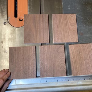 Könnte beinhalten: Fünf quadratische Holzblöcke mit einer satten braunen Farbe und sichtbarer Holzmaserung sind auf einer Metalloberfläche angeordnet. Die Blöcke sind wahrscheinlich aus Walnussholz oder einem ähnlichen Hartholz geschnitten. Eine Hand ist sichtbar, die einen der Blöcke berührt.