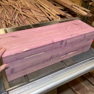 Può includere: Una scatola rettangolare in legno di cedro non rifinito con una tonalità rosa-viola, appoggiata su una sega da tavolo in metallo. La venatura del legno è visibile. La scatola è circondata da altri pezzi di legno.