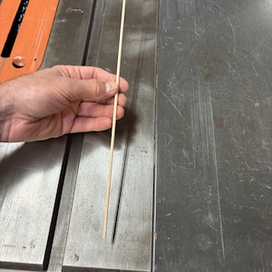 Puede incluir: Una varilla de madera delgada de color claro sostenida verticalmente. La varilla está colocada contra una superficie de mesa de metal con una regla visible en el borde inferior, mostrando que la longitud supera la marca de 25 cm.