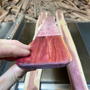 May include: A piece of unfinished wood, showcasing a blend of reddish-brown and light purple hues. The wood grain is visible, and the piece is held by a hand, suggesting a woodworking project. The background includes other wood pieces and a saw.