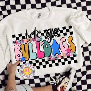 May include: White sweatshirt with the words "let's go BULLDOGS" in colorful, bubble letters. The design includes stars and a checkered pattern. A person is wearing the sweatshirt with denim shorts and sneakers.