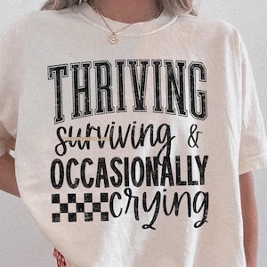 May include: White t-shirt with black text that reads "THRIVING surviving & OCCASIONALLY crying" with a checkered pattern below the text.