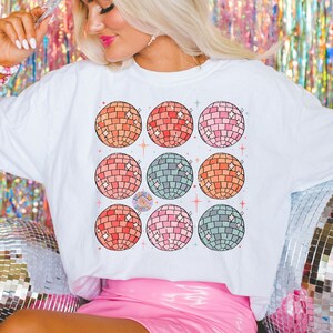 May include: A white t-shirt with a design of nine colorful disco balls. The shirt is worn by a person wearing a pink cowboy hat and a pink skirt. The background is a shimmering silver curtain.