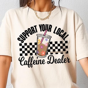 May include: A beige t-shirt with a black and white checkered design and the text "Support Your Local Caffeine Dealer" with a cartoon image of a cup of iced coffee with a pink straw and flowers.