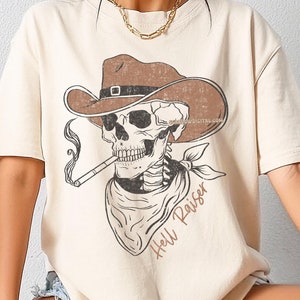 May include: A beige t-shirt with a graphic of a skeleton wearing a cowboy hat and bandana. The text "Hell Raiser" is printed below the graphic.
