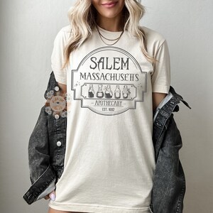 Może przedstawiać: Beżowa koszulka z nadrukiem graficznym vintage'owego szyldu apteki z napisem "Salem, Massachusetts - Apothecary - Est. 1692".