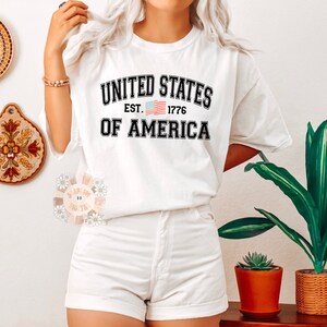 May include: White t-shirt with black text that reads "United States Est. 1776 Of America" with a small American flag graphic.