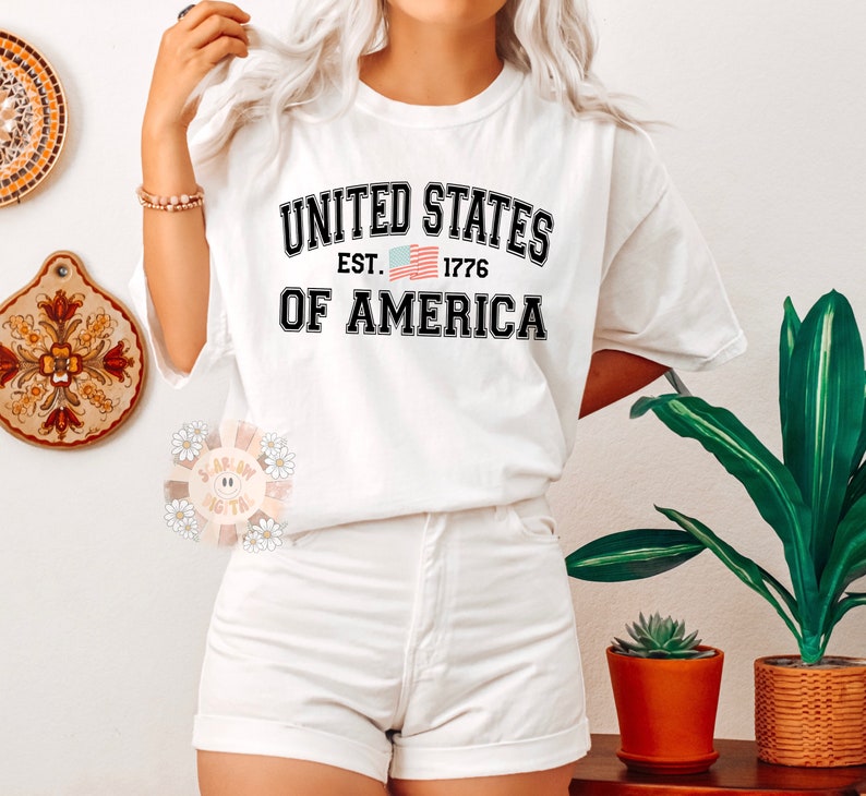 May include: White t-shirt with black text that reads "United States Est. 1776 Of America" with a small American flag graphic.