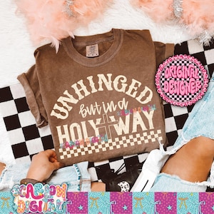 Puede incluir: Una camiseta marrón con las palabras "UNHINGED but in a HOLY WAY" en una fuente vintage. La camiseta tiene un gráfico de cruz y un patrón de cuadros. La camiseta se muestra sobre una superficie a cuadros en blanco y negro.