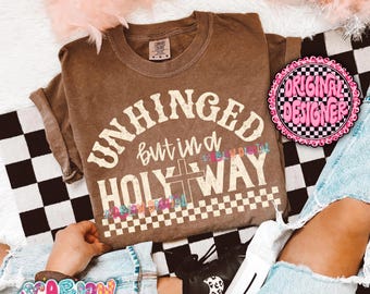 May include: A brown t-shirt with the words "UNHINGED but in a HOLY WAY" in a vintage font. The shirt has a cross graphic and a checkered pattern. The shirt is displayed on a black and white checkered surface.