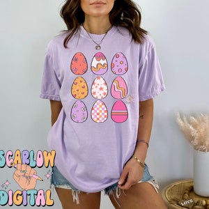 May include: A purple t-shirt with a design of nine colorful Easter eggs. Each egg has a different pattern, including hearts, stars, flowers, and stripes.
