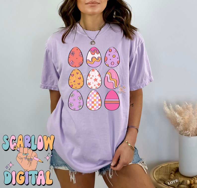 May include: A purple t-shirt with a design of nine colorful Easter eggs. Each egg has a different pattern, including hearts, stars, flowers, and stripes.