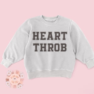 Op de afbeelding: Een lichtgrijs sweatshirt met de tekst "HEART THROB" bedrukt in zwart met een witte omtrek.