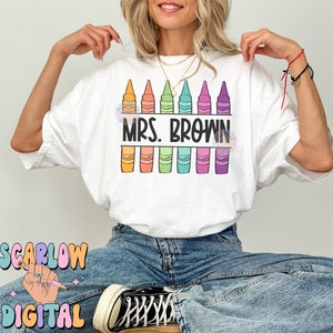 以下が含まれることがあります： 白いTシャツに、カラフルなクレヨンが並んだデザインと「Mrs. Brown」の文字がプリントされています。