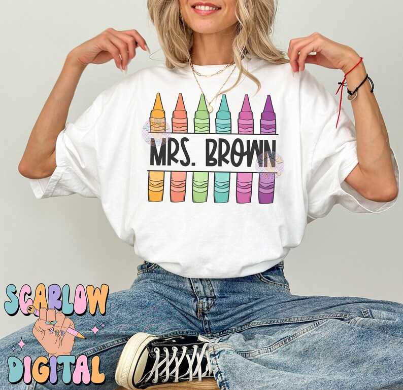 以下が含まれることがあります： 白いTシャツに、カラフルなクレヨンが並んだデザインと「Mrs. Brown」の文字がプリントされています。