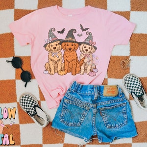 K&ouml;nnte beinhalten: Ein pinkfarbenes T-Shirt mit einem Aufdruck von drei Golden Retrievern, die Hexenh&uuml;te tragen und K&uuml;rbisse halten. Das T-Shirt liegt auf einer karierten Oberfl&auml;che mit einer schwarzen Sonnenbrille und einem Paar karierten Vans-Schuhen.