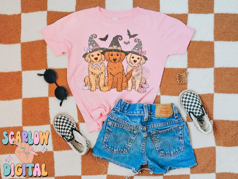 K&ouml;nnte beinhalten: Ein pinkfarbenes T-Shirt mit einem Aufdruck von drei Golden Retrievern, die Hexenh&uuml;te tragen und K&uuml;rbisse halten. Das T-Shirt liegt auf einer karierten Oberfl&auml;che mit einer schwarzen Sonnenbrille und einem Paar karierten Vans-Schuhen.