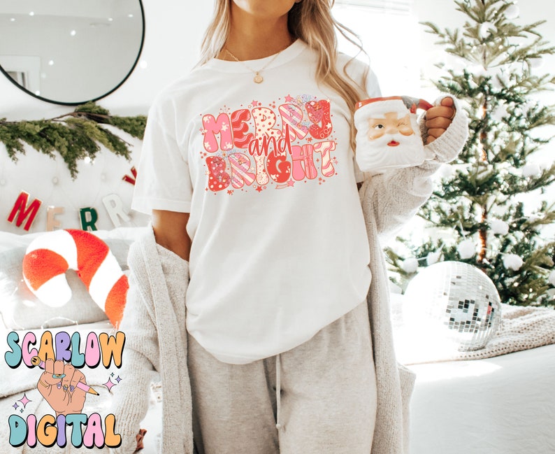 Op de afbeelding: Wit T-shirt met de woorden "Merry and Bright" in een kleurrijk retro-lettertype. Het ontwerp bevat roze, rode en witte elementen. Een Santa-mok wordt in de hand gehouden. Het algemene thema is Kerstmis.