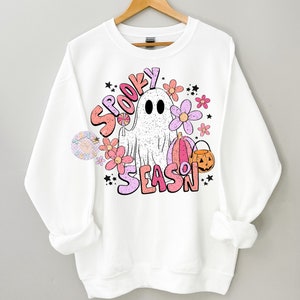 Pu&ograve; includere: Felpa bianca con un design grafico rosa, arancione e bianco di un fantasma, fiori, zucche e il testo "Spooky Season".